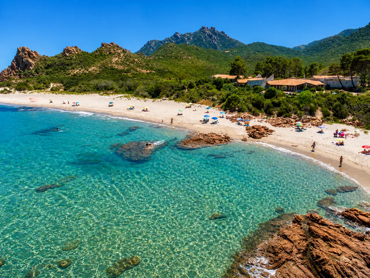 Marina di Gairo — baia con sabbia chiara, rocce di porfido rosso e mare turchese sotto i monti